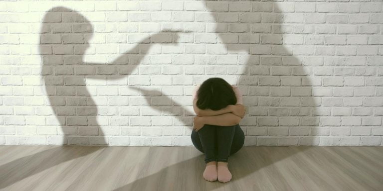 Imagen de una mujer sentada en el suelo con la cabeza gacha y en la pared unas sombras de un hombre y una mujer discutiendo. Escena que representando la violencia machista.